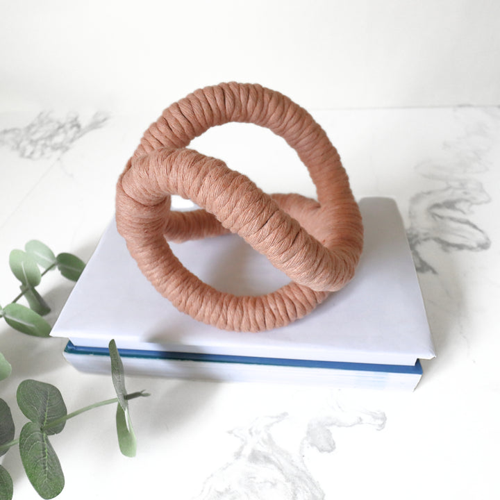 Close up of peach color desk accessory displayed on books showcasing intricate hand knotted details of infinity rope sculpture, adding artistic flair to a modern interior.